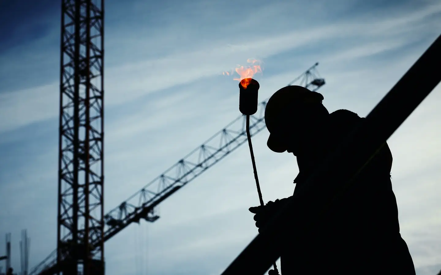 Silhouette of a worker holding a lit gas torch beneath a tower crane